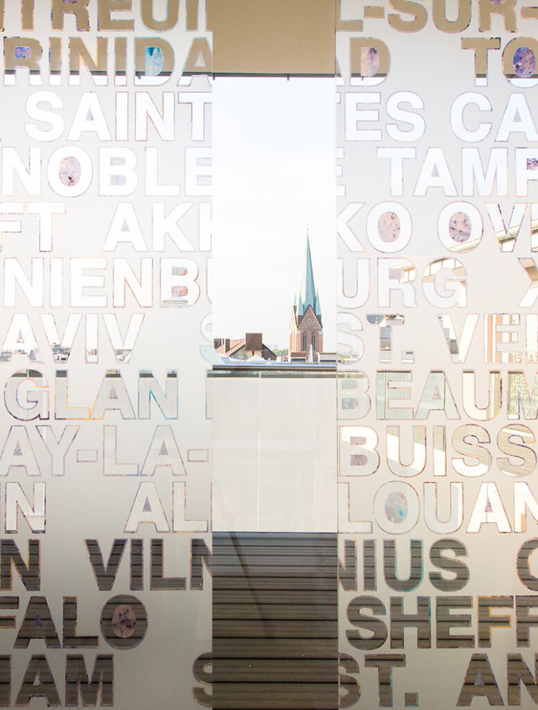 Text auf Fenster, reflektiert Licht. Kirchenspitze und Architekturelement im Hintergrund sichtbar.