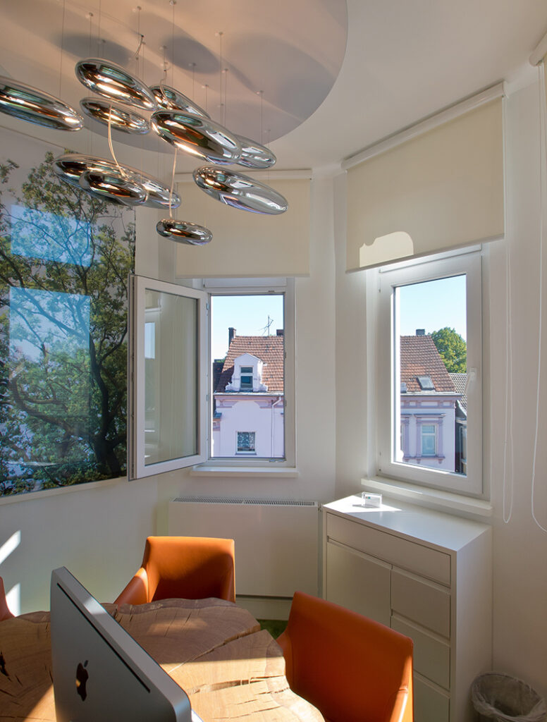 Ein modernes Büro mit hellen Wänden, Baumfensterbild, orangen Stühlen und geschwungener Deckenleuchte.