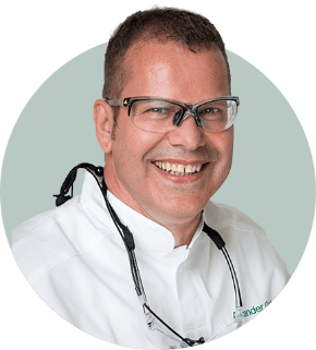 Mann mit Brille lächelt, trägt weißen Kittel, schwarzes Brillenband und Schutzbrille.
