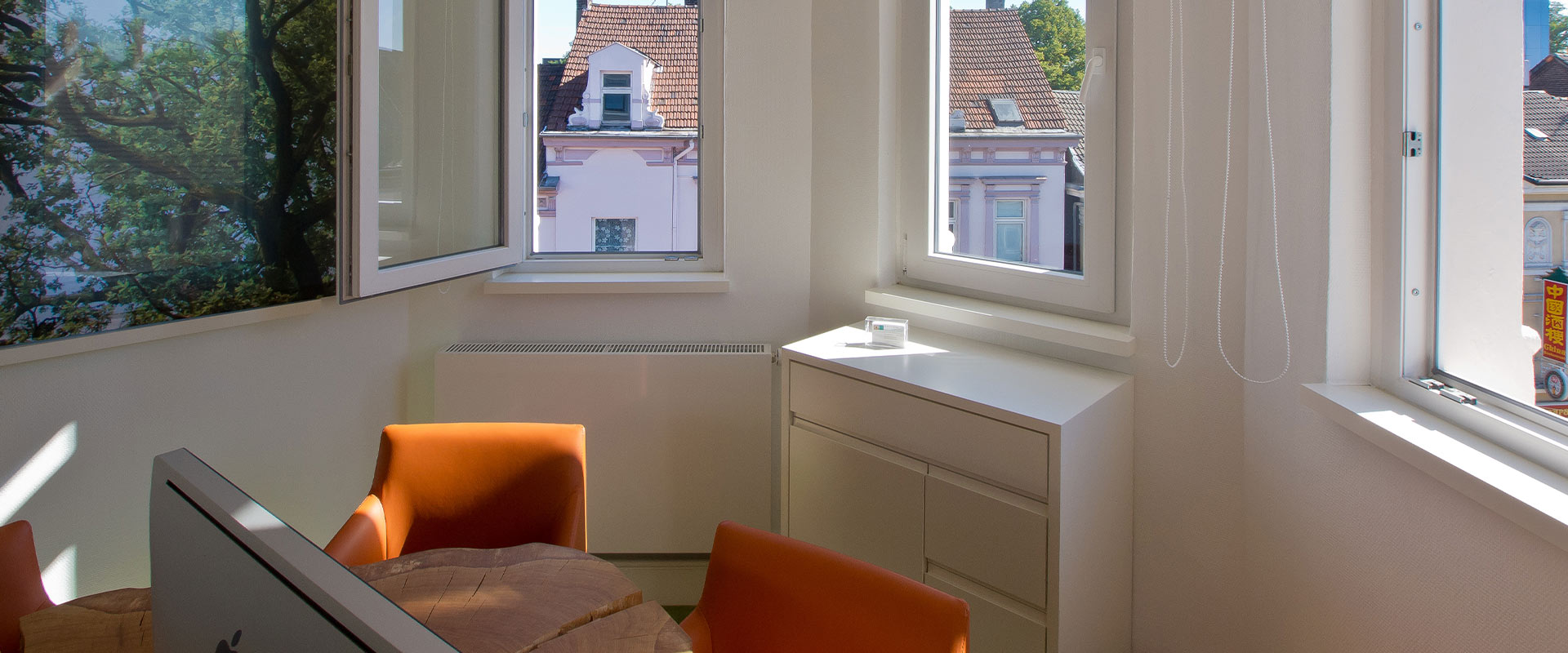 Helles Büro mit offenen Fenstern, orangenen Stühlen und Blick auf rosa Häuser im Sonnenlicht.