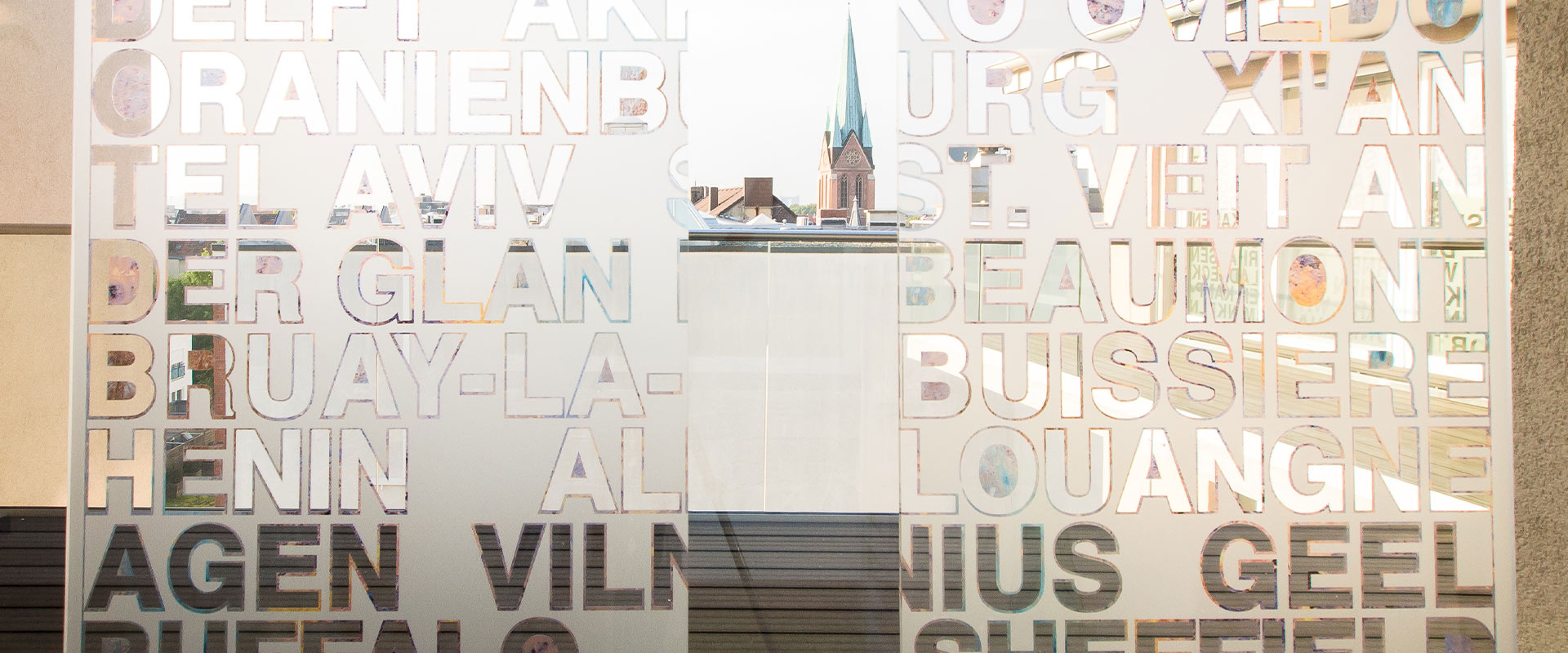 Eine durchscheinende Wand mit Städtenamen in Großbuchstaben vor einer Stadtansicht.