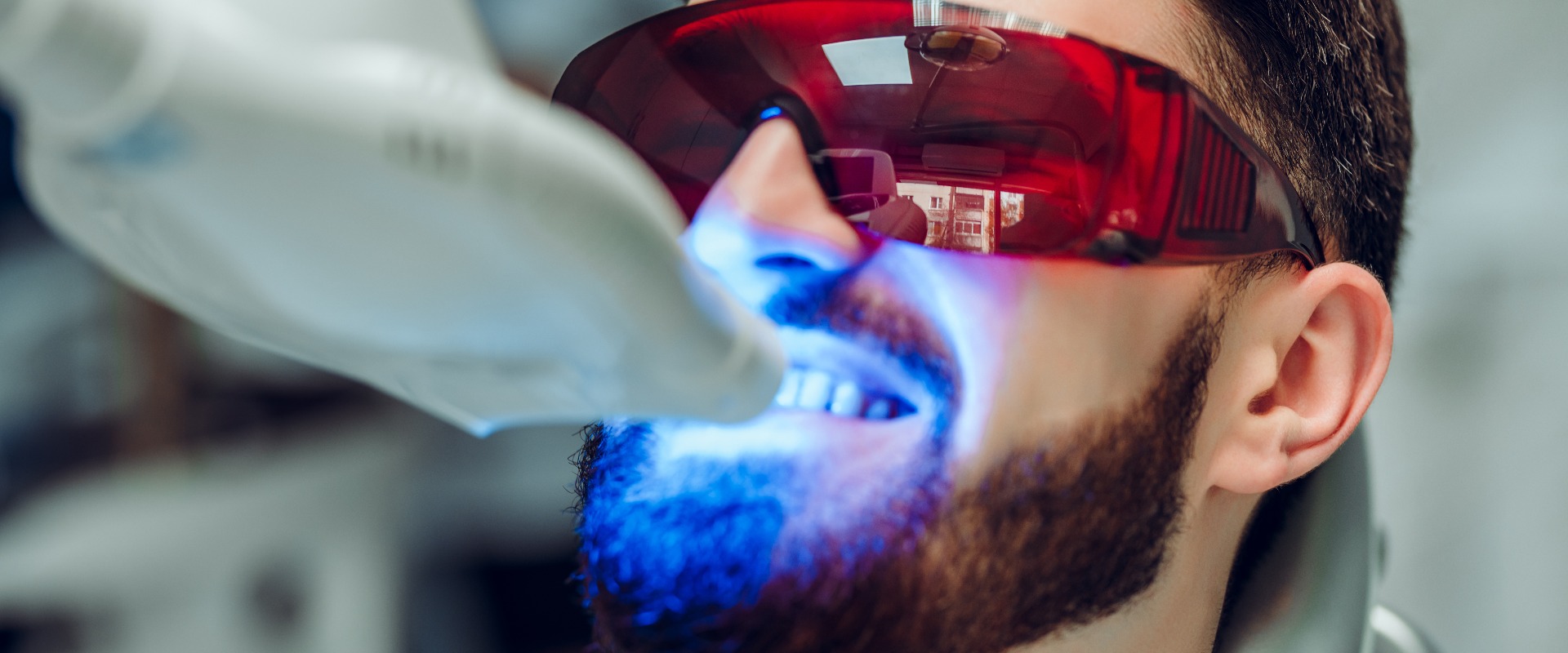 Mann bei einer Zahnaufhellung mit einem zahnärztlichen UV-Bleichgerät, Zahnarzthelferin kümmert sich um den Patienten, Augen mit Brille geschützt. Bleaching-Behandlung mit Licht, Laser und Fluorid.