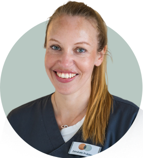 Eine lächelnde Frau im blauen Kittel mit Namensschild und hellbraunen Haaren